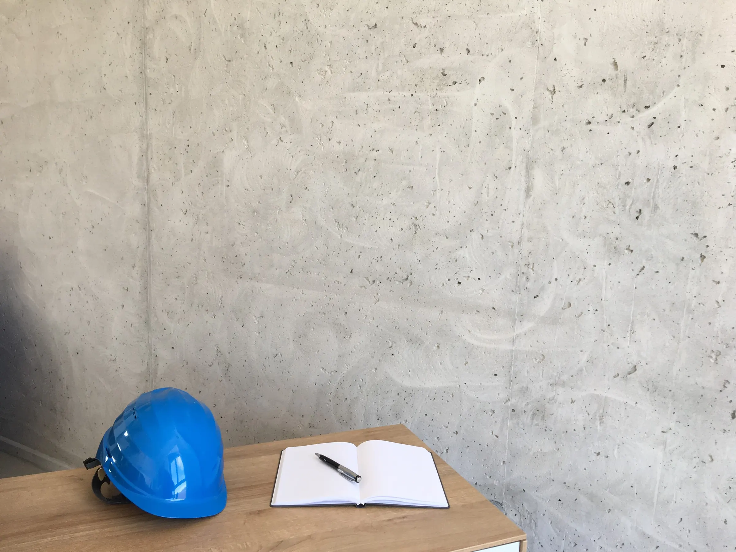Blauer Bauhelm und Notizbuch vor einer Sichtbetonwand
